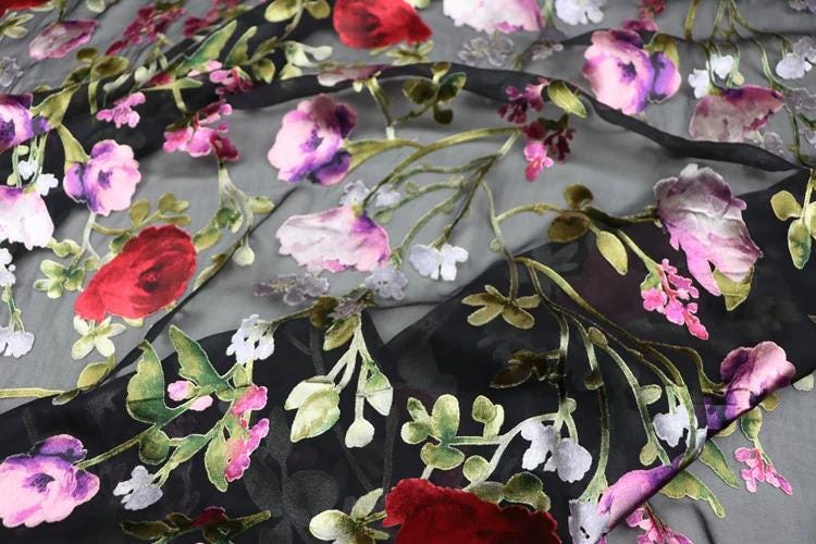 a close-up view of a fabric with a floral pattern. The fabric is predominantly black, with red and white flowers and leaves scattered across it. The flowers appear to be in various stages of bloom, creating a vibrant and lively visual effect. The fabric is described as sheer, which suggests that it has a lightweight and flowing texture.