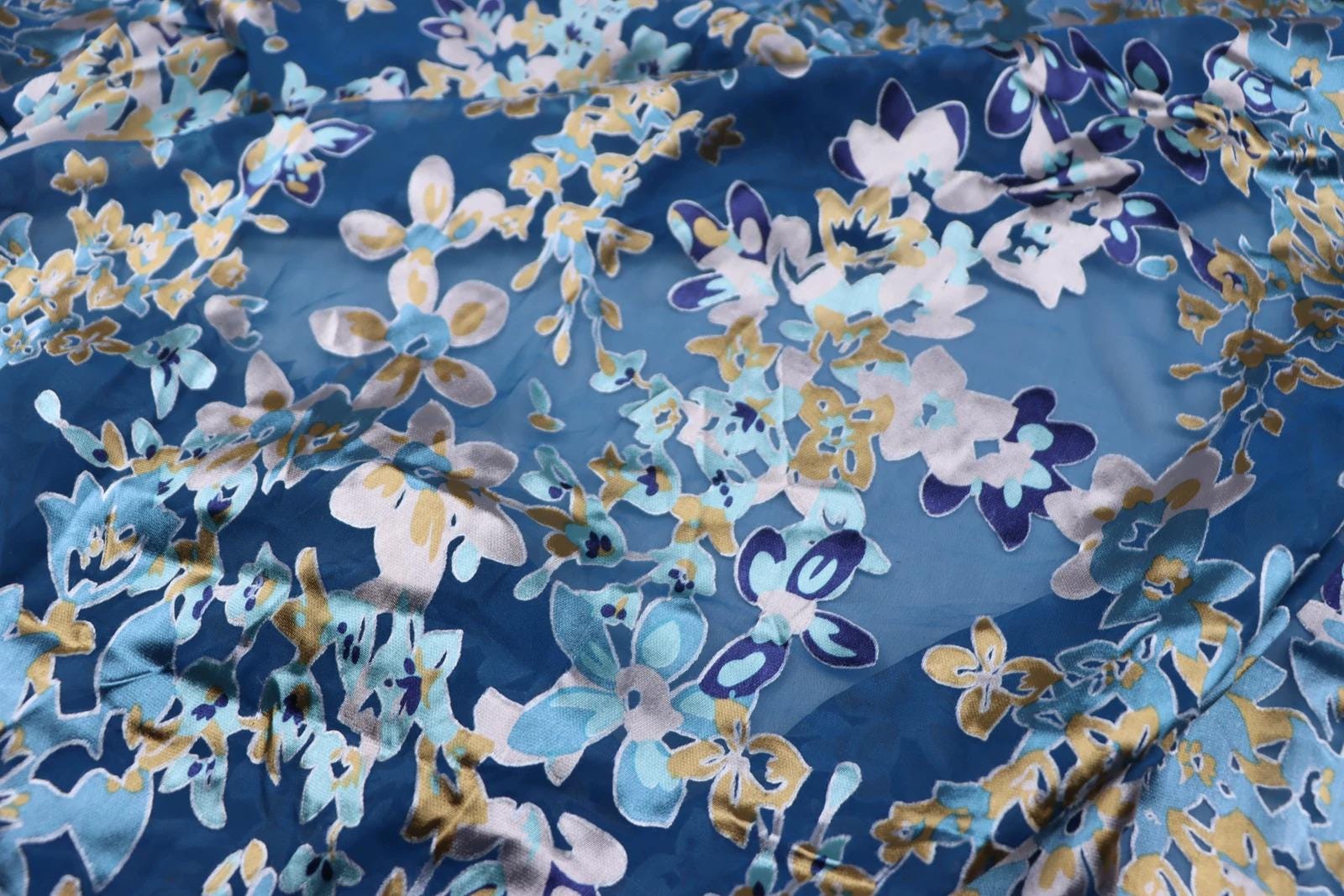 a close-up view of a fabric with a floral pattern in shades of blue, white, and gold. The pattern consists of various flowers and leaves, creating a visually appealing and intricate design.