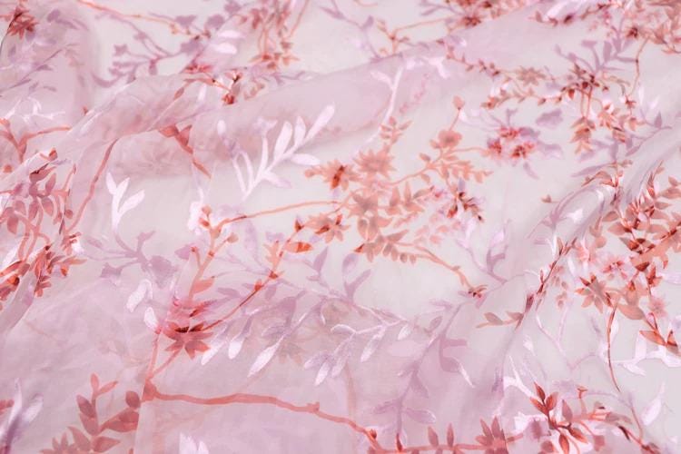 a close-up view of a fabric with a pink and red floral pattern. The fabric appears to be made of sheer material, giving it a delicate and airy appearance. The pattern consists of various shades of pink and red flowers and leaves, creating a visually appealing and intricate design.