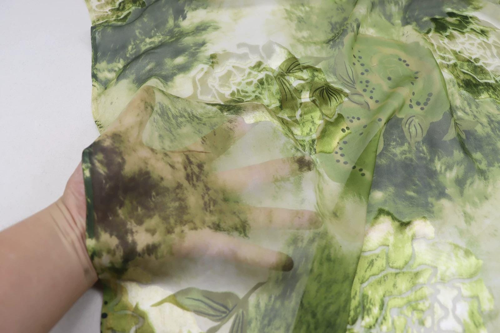 A hand is holding a piece of fabric with a green and white floral pattern, which appears to be a hand-made or handmade textile.
