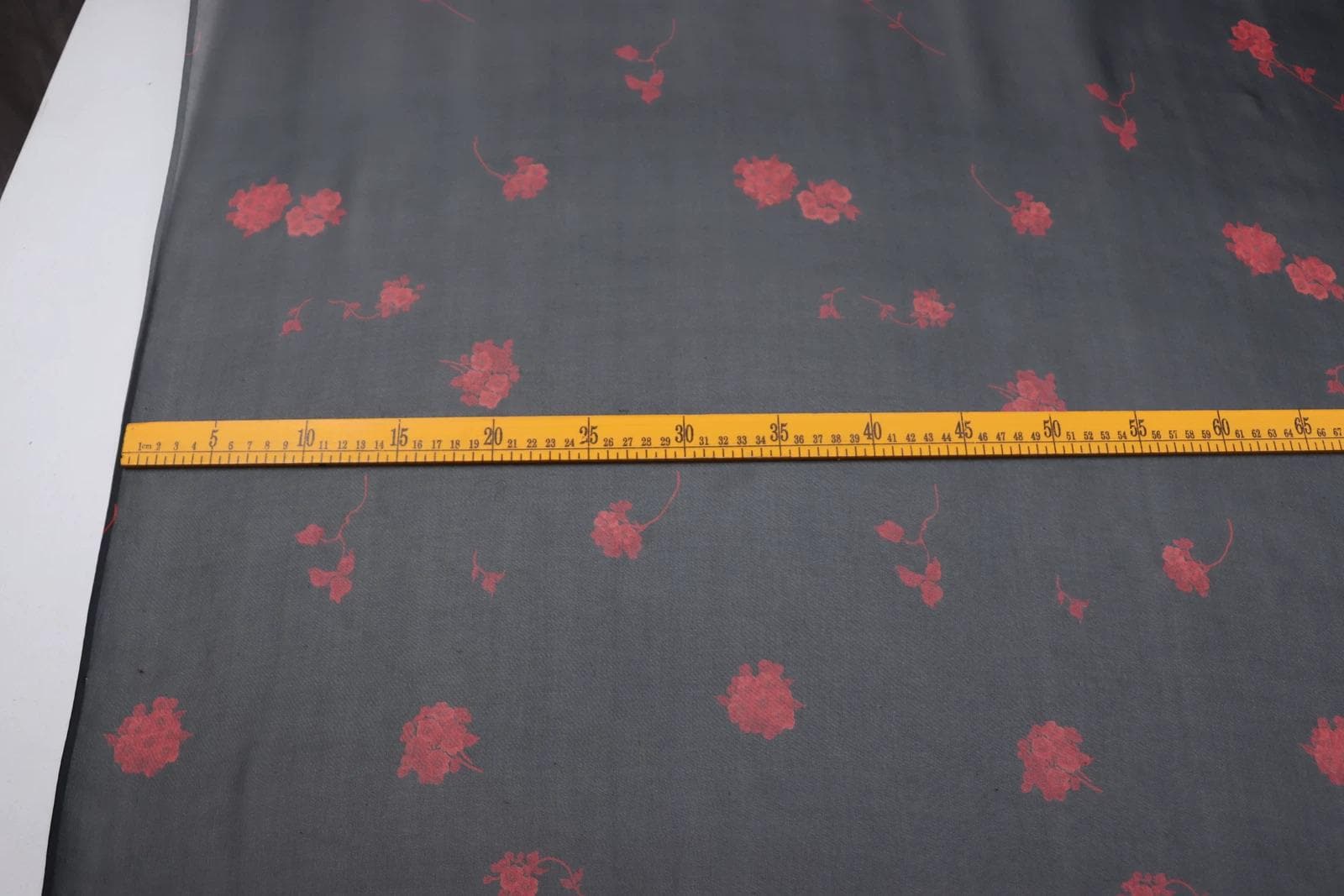 a close-up view of a fabric with a floral pattern, featuring red flowers on a black background. A yellow measuring tape is laid across the fabric, indicating the size or dimensions of the fabric.