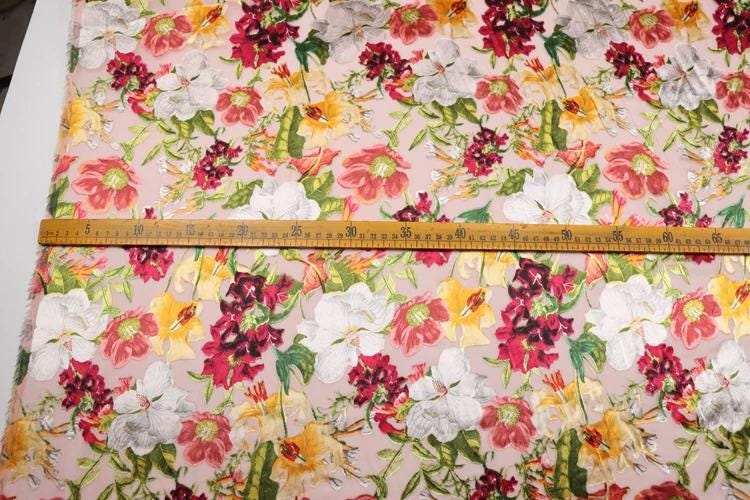 a fabric with a floral pattern, featuring various colors and types of flowers. A ruler is placed on the fabric, indicating the size or dimensions of the pattern.