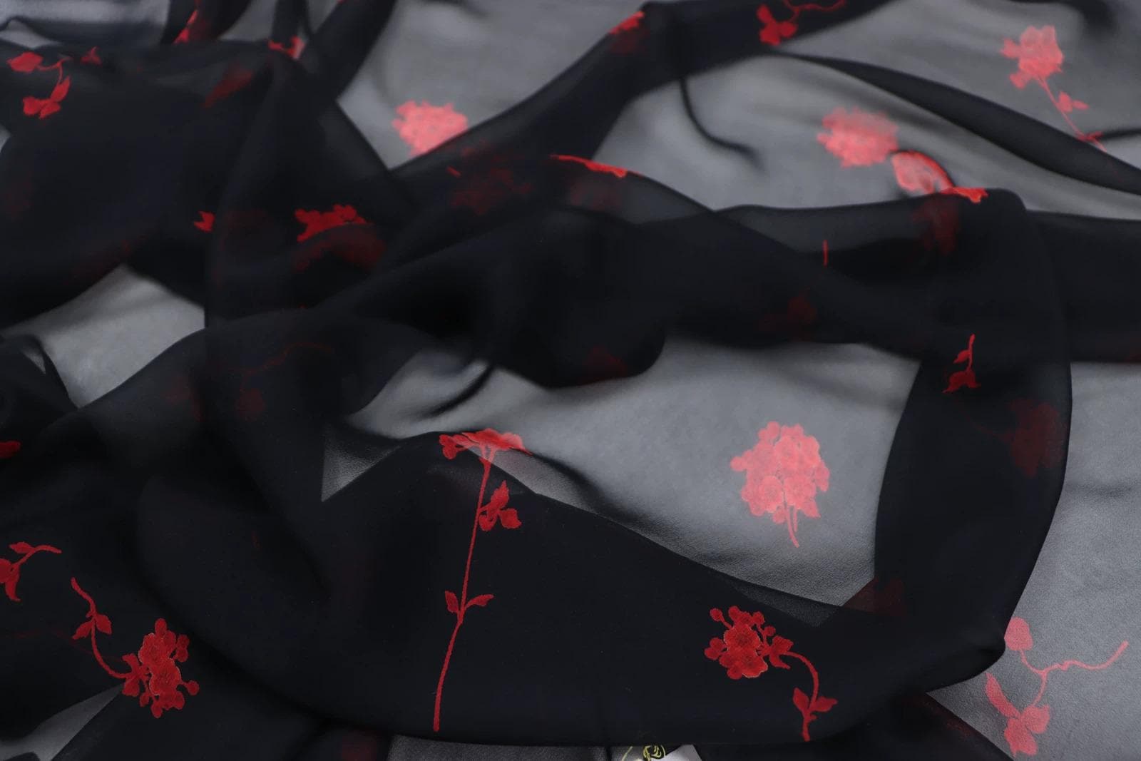 a close-up view of a black fabric with red floral patterns. The fabric appears to be made of silk or chiffon, and the red flowers are scattered across the fabric in a random pattern.