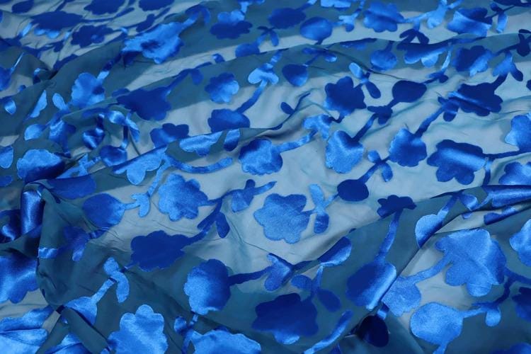 a close-up view of a fabric with a blue floral pattern. The fabric appears to be made of a shiny, silky material and has a crumpled texture. The pattern consists of blue flowers or leaves, which are the main focus of the image. The fabric is spread out, covering a significant portion of the frame, and the close-up perspective allows for a detailed view of the intricate design.