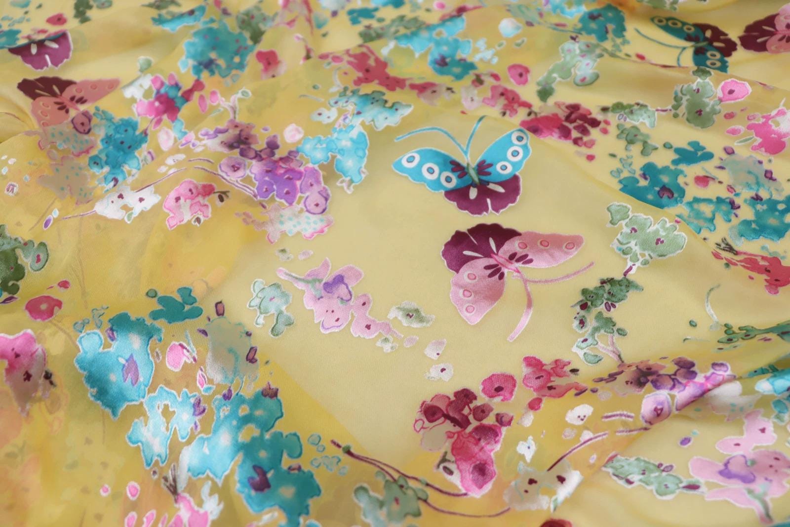 The image is a close-up of a fabric with a vibrant, colorful floral pattern. The fabric appears to be made of silk or a similar material, and it features a variety of flowers and butterflies in shades of pink, blue, green, and yellow. The pattern is intricate and detailed, with each flower and butterfly adding to the overall visual appeal of the fabric.