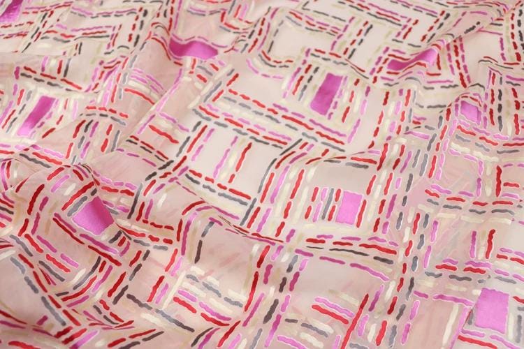 a close-up view of a fabric with a geometric pattern in shades of pink, red, and gray. The pattern consists of various shapes and lines, creating a visually striking and intricate design.