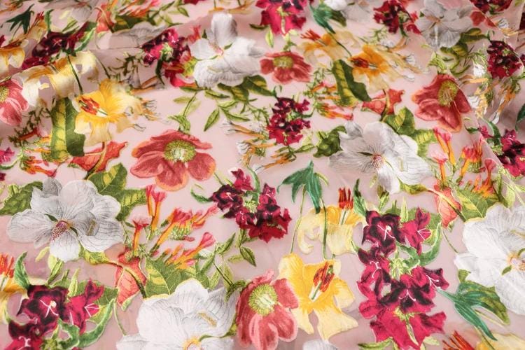 The image is a close-up view of a fabric with a vibrant floral pattern. The fabric is adorned with a variety of colorful flowers, including red, white, yellow, and green, arranged in a visually appealing manner. The flowers are depicted in different sizes and orientations, creating a dynamic and lively appearance. The fabric appears to be made of a soft, delicate material, suitable for clothing or other textile applications.