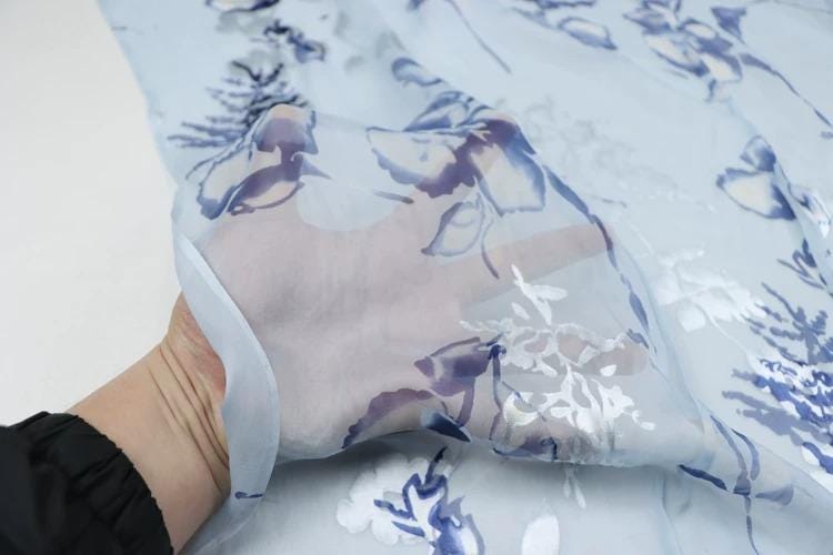 A hand is holding a piece of fabric with a floral pattern, which appears to be a light blue material.