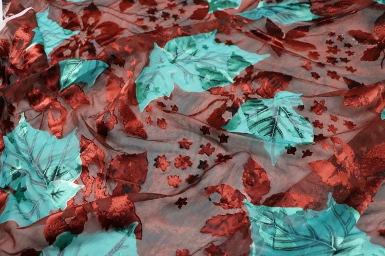 a close-up view of a fabric with a vibrant pattern of red and green leaves and flowers. The fabric appears to be made of sheer material, allowing the leaves and flowers to be clearly visible. The pattern is intricate and detailed, creating a visually striking and textured appearance.