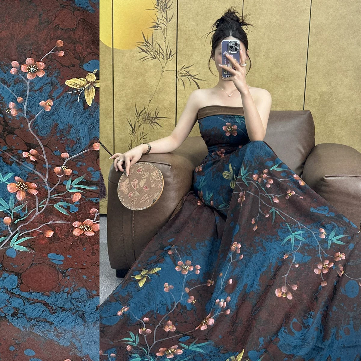 A woman in a long, floral-patterned dress is sitting on a leather chair, holding a fan and taking a selfie with her phone.