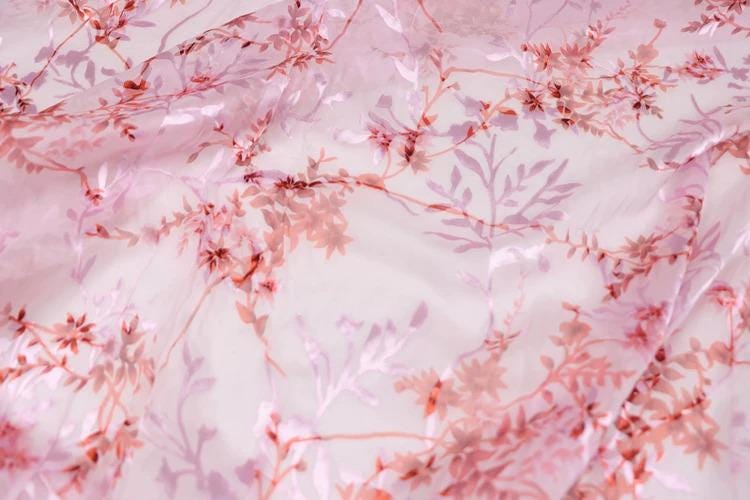 a close-up view of a fabric with a pink floral pattern. The fabric appears to be made of sheer material, with delicate pink flowers and leaves covering the entire surface. The pattern is intricate and detailed, creating a visually appealing and textured appearance.