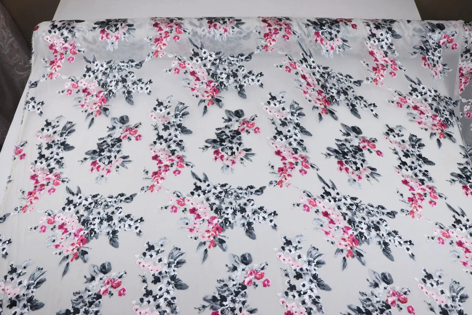 a fabric with a floral pattern in shades of pink and black, with some white flowers interspersed throughout. The pattern is repeated across the fabric, creating a visually appealing and intricate design.