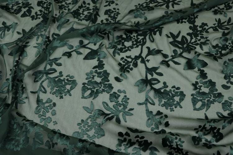 a close-up view of a fabric with a floral pattern, featuring green leaves and flowers. The fabric appears to be made of a sheer material, giving it a delicate and airy appearance.