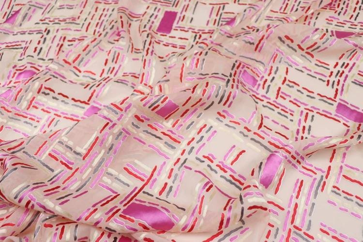 a close-up view of a fabric with a geometric pattern in shades of pink, red, and gray. The pattern consists of intersecting lines and rectangles in various sizes and orientations, creating a visually striking and intricate design.