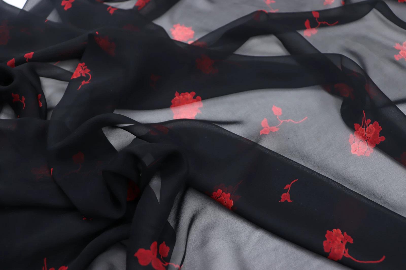 a close-up view of a black fabric with red floral patterns. The fabric appears to be made of sheer material, giving it a delicate and flowing appearance. The red flowers are scattered across the fabric, creating a visually striking contrast against the black background.