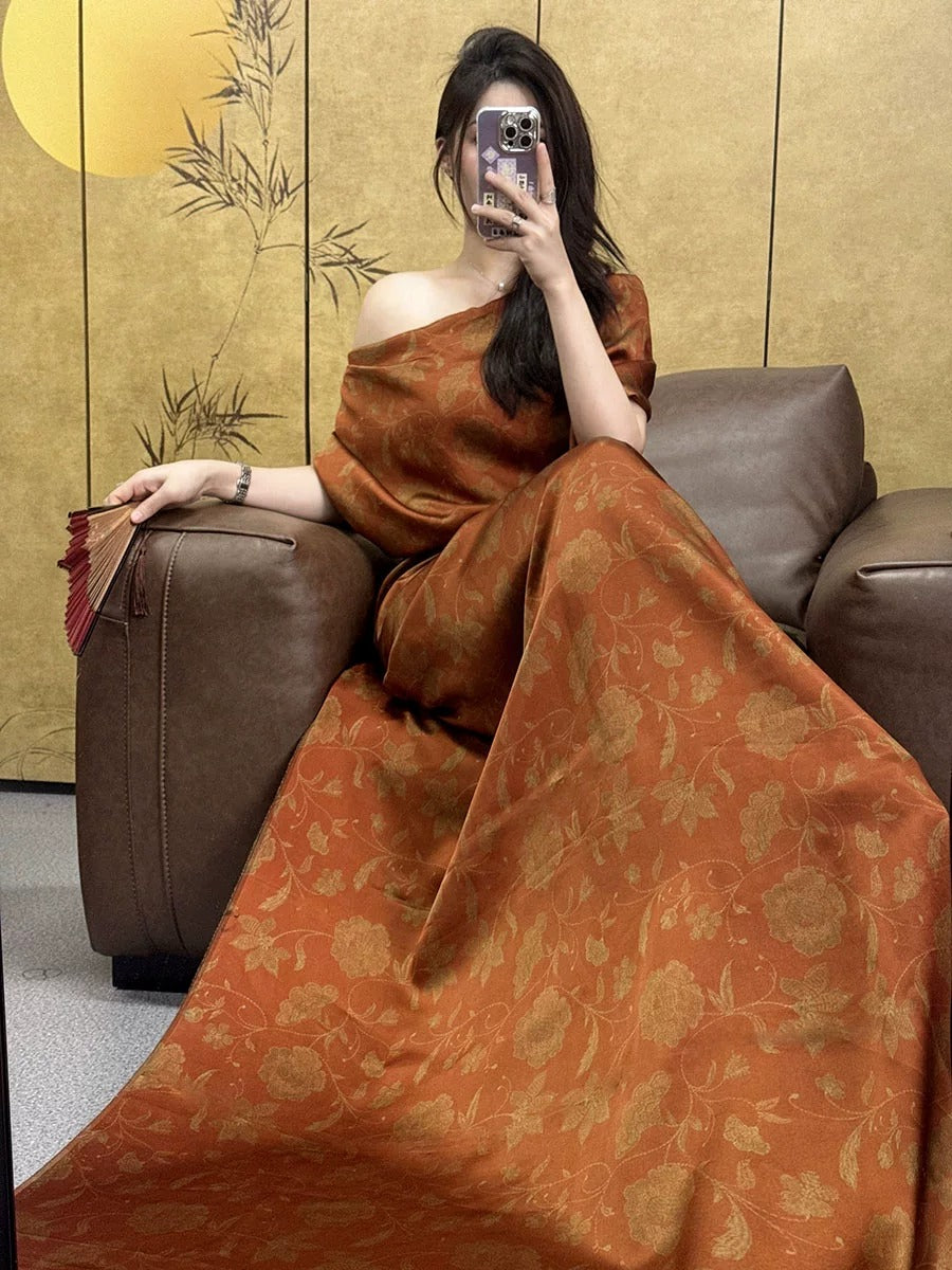 A woman in an orange dress is sitting on a brown leather couch, holding a fan and taking a selfie with her phone.