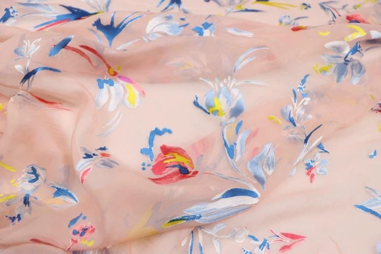 a close-up view of a fabric with a floral pattern in various colors, including pink, blue, and yellow. The fabric appears to be made of silk or chiffon, and the pattern is intricate and detailed.