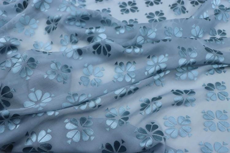 a close-up view of a fabric with a repeating pattern of blue and white flowers. The fabric appears to be made of a soft, flowing material, and the pattern is intricate and detailed.