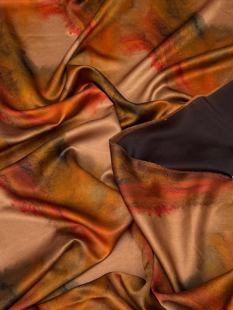 The image is a close-up of a fabric with a vibrant, abstract pattern in shades of orange, brown, and black. The fabric appears to be made of silk or a similar material, and it is crumpled, giving it a textured and dynamic appearance.