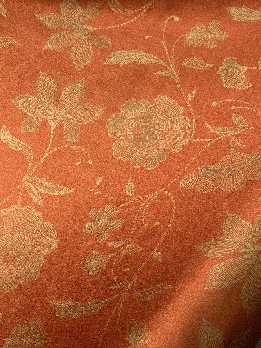 a close-up view of a fabric with a floral pattern in shades of orange and gold. The pattern features large, detailed flowers and leaves, creating a visually striking and intricate design.