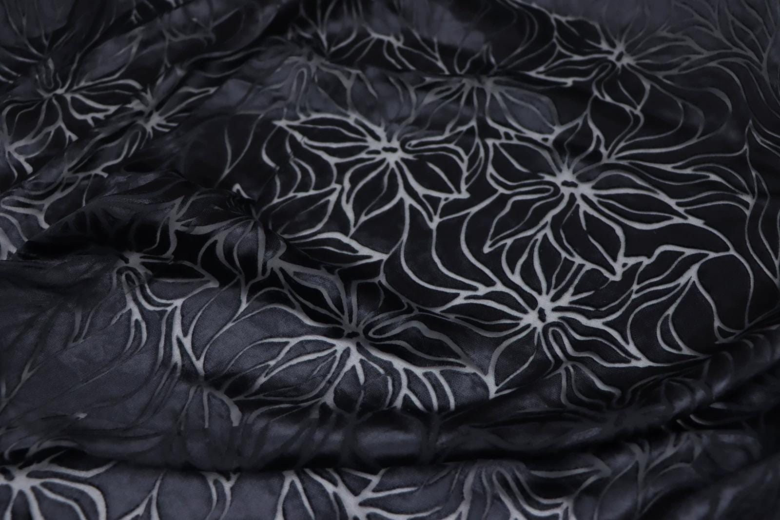 a close-up view of a fabric with a black and white floral pattern. The fabric appears to be made of a shiny material, possibly silk or satin, and has a textured appearance. The pattern consists of stylized flowers or leaves, with some areas appearing more detailed and intricate than others. The overall composition of the image focuses on the fabric's texture and the repeating floral pattern.
