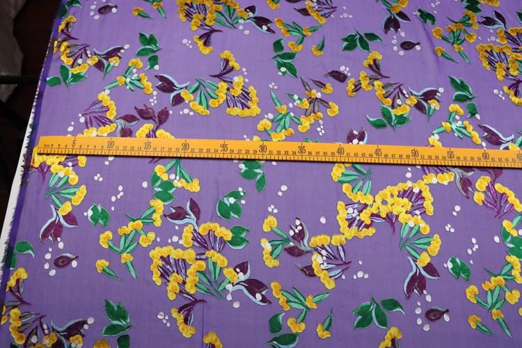 a fabric with a vibrant purple background adorned with yellow and green floral patterns. A yellow measuring tape is laid across the fabric, providing a size reference.