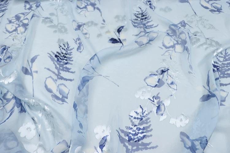 a close-up view of a fabric with a floral pattern in shades of blue and white. The fabric appears to be made of sheer material, giving it a delicate and airy appearance. The pattern consists of various floral designs, including leaves, flowers, and branches, which are spread across the fabric in a visually appealing manner.