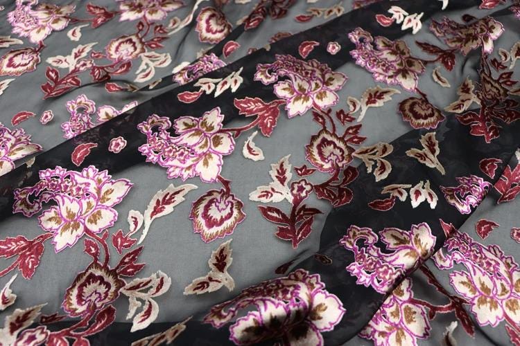 a close-up view of a fabric with a floral pattern in shades of pink, red, and black. The fabric appears to be a sheer material, with a dark background that contrasts with the lighter colors of the flowers. The pattern is intricate and detailed, with the flowers and leaves filling the entire visible area of the fabric.