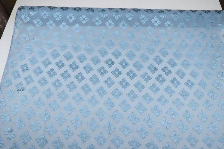 a close-up view of a fabric with a repeating pattern of blue flowers or leaves. The pattern is intricate and detailed, covering the entire surface of the fabric. The fabric appears to be a type of textile or material, possibly used for clothing or home decor.