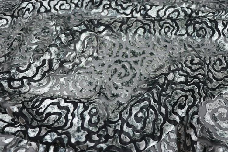 a close-up view of a fabric with a repeating pattern of black and white swirls or curves. The pattern is intricate and detailed, creating a visually striking and textured appearance.