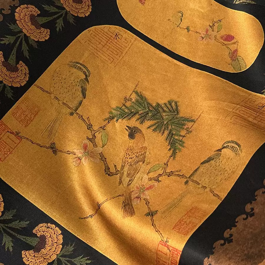 a close-up view of a fabric with a detailed, hand-painted design of birds perched on branches and flowers. The design is predominantly gold in color, with accents of black and brown. The fabric appears to be a piece of clothing or textile, with the intricate design adding a unique and artistic touch to the overall appearance.