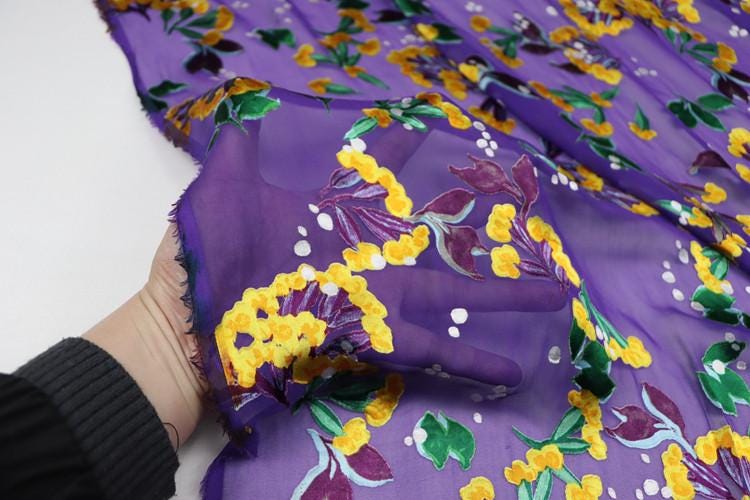 A hand is holding a piece of fabric with a vibrant purple and yellow floral pattern.