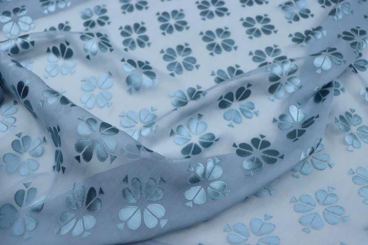 a close-up view of a fabric with a repeating pattern of blue and white flowers or leaves. The fabric appears to be made of a soft, flowing material, and the pattern is intricate and detailed.