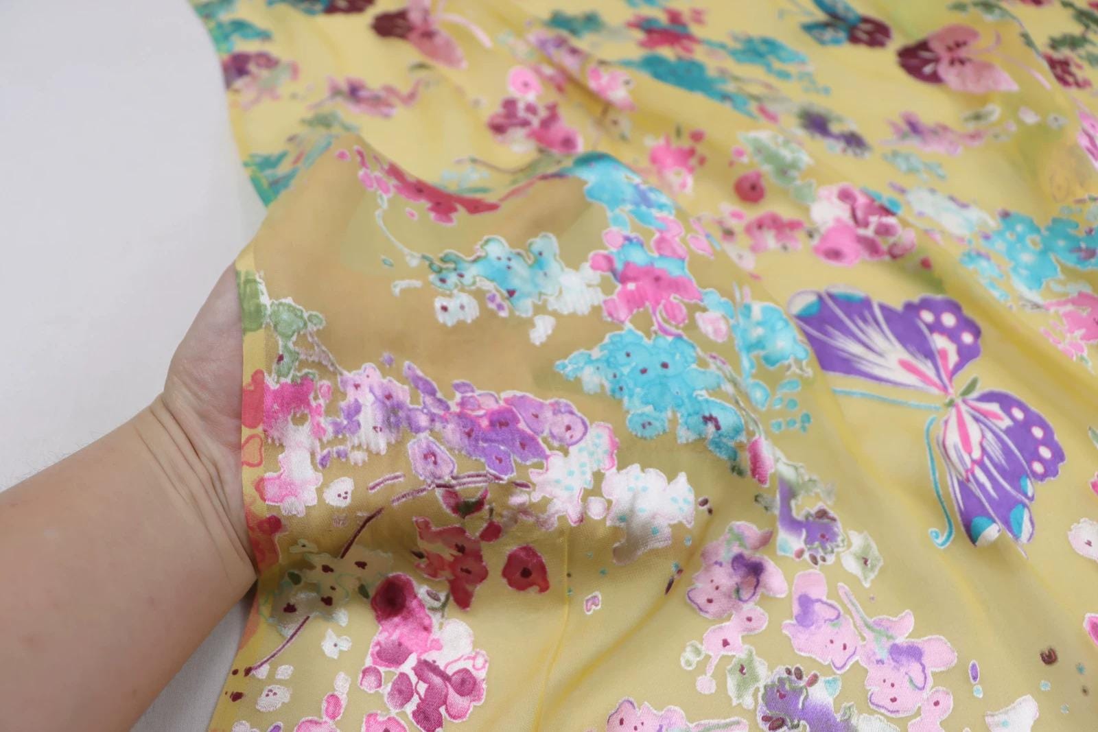 A hand is holding up a fabric with a vibrant, colorful floral pattern, including a butterfly design.