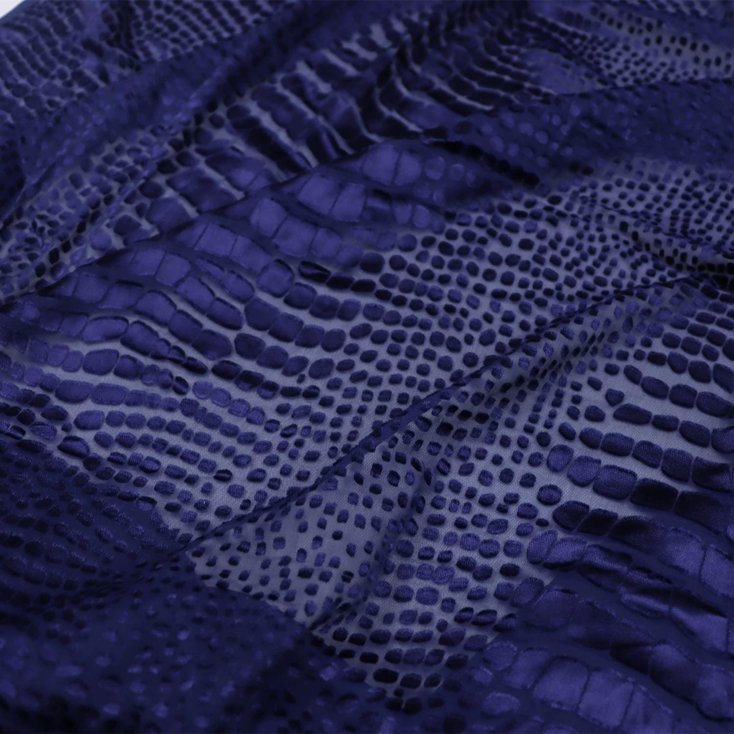 a close-up view of a fabric with a distinctive pattern of small dots or circles, resembling the scales of a snake or a lizard. The fabric appears to be a dark blue or purple color, with a textured and wrinkled appearance.