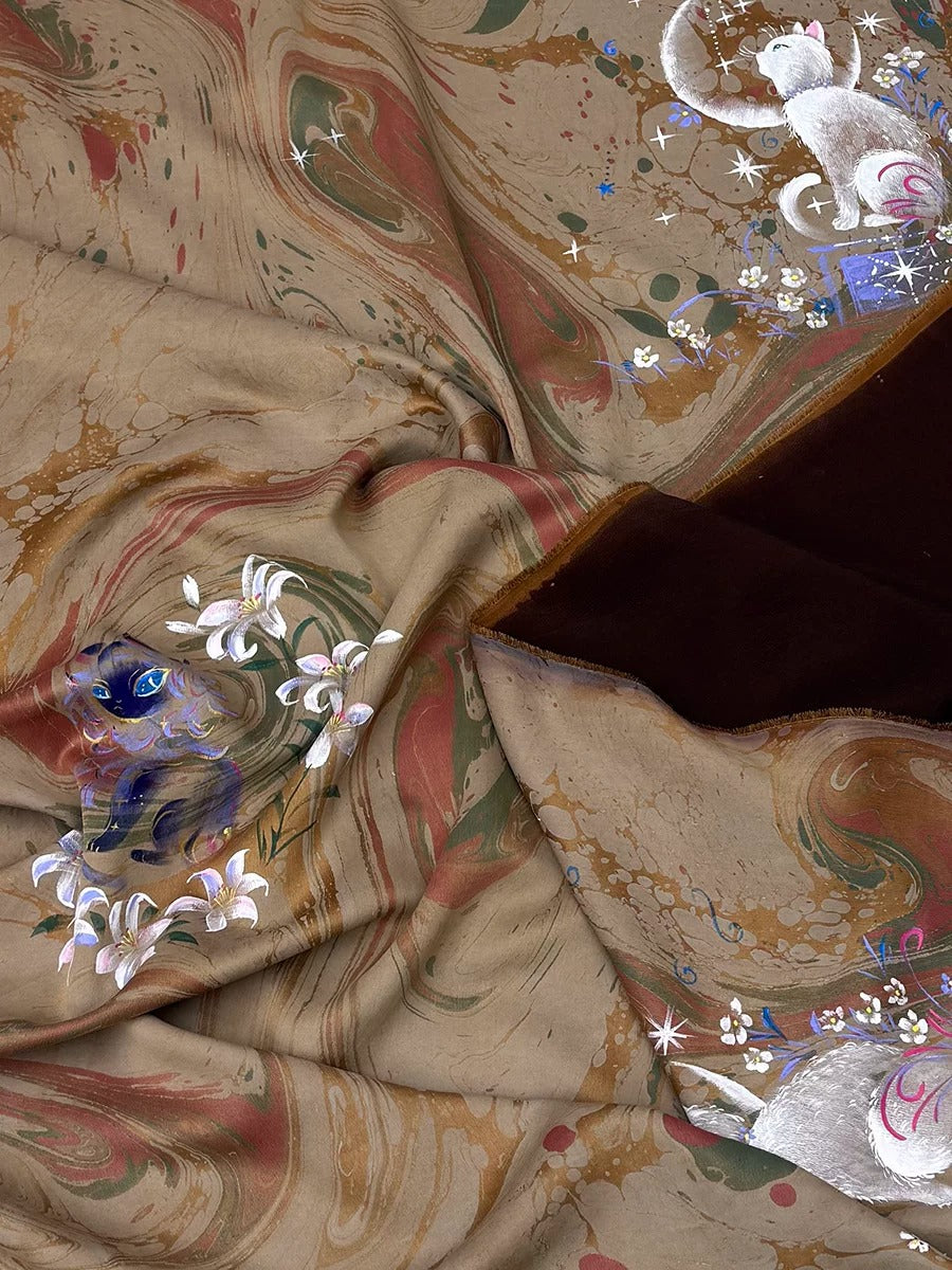 The image is a close-up of a fabric with a marbled pattern, featuring a mix of brown, green, and white colors. The fabric appears to be made of silk, and there are small white flowers and a blue and white cat design visible on the fabric.