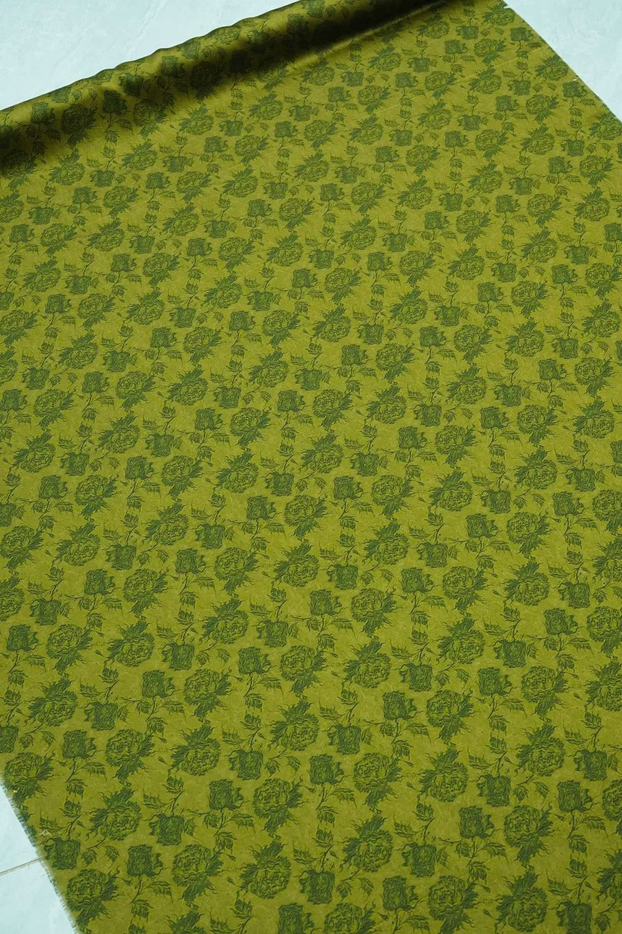 a close-up view of a fabric with a repeating pattern of green floral designs. The pattern is intricate and detailed, covering the entire surface of the fabric. The fabric appears to be a type of textile, possibly used for clothing or home decor.