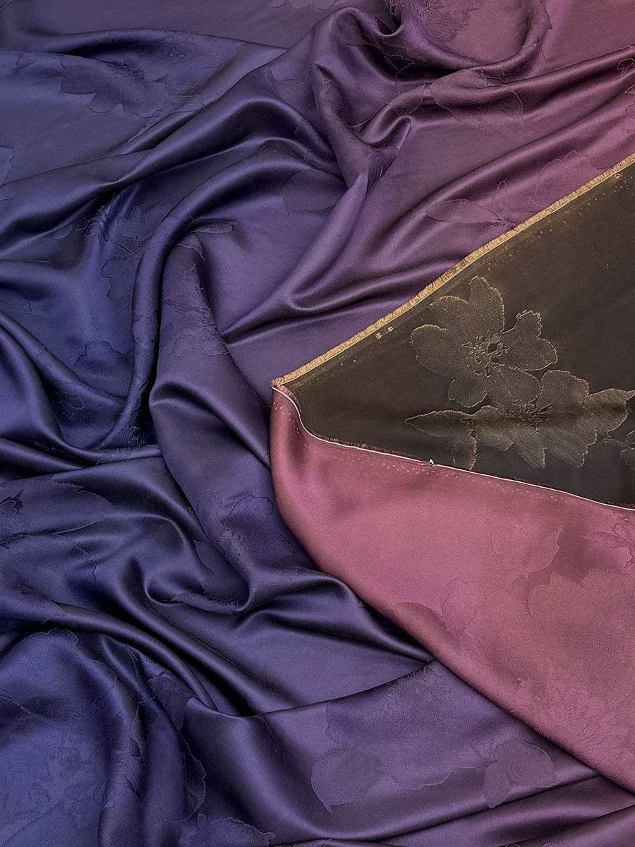 The image is a close-up of a fabric with a floral pattern, featuring a mix of purple and pink colors. The fabric appears to be made of silk or satin, with a crinkled texture that gives it a soft and luxurious feel.