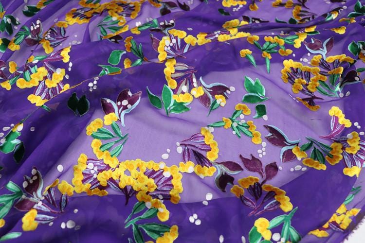 a close-up view of a fabric with a vibrant floral pattern in shades of purple and yellow. The pattern features various flowers and leaves, creating a visually striking and colorful design.