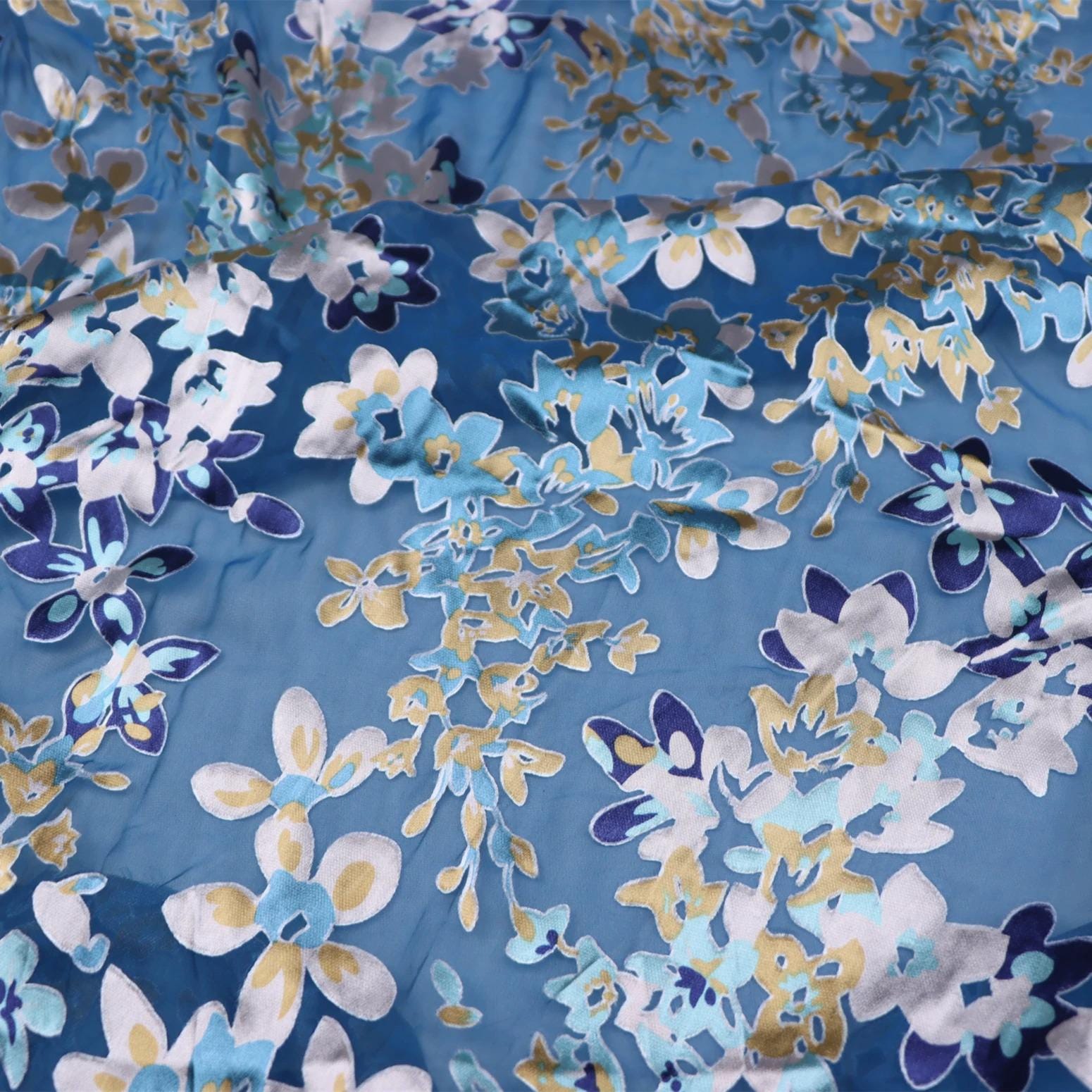 a close-up view of a fabric with a floral pattern in shades of blue, white, and gold. The pattern consists of various flower shapes and sizes, creating a visually appealing and intricate design.