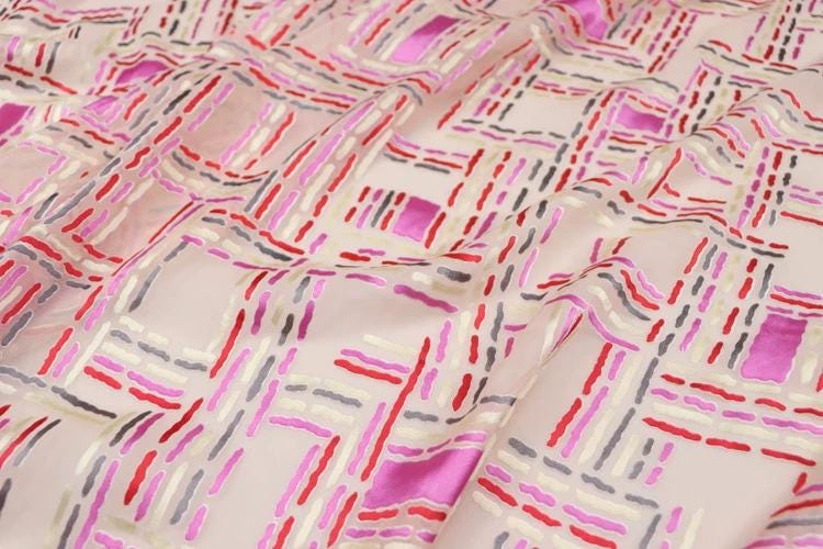 a close-up view of a fabric with a geometric pattern in shades of pink, red, and gray. The pattern consists of various shapes and lines, creating a visually striking and intricate design.