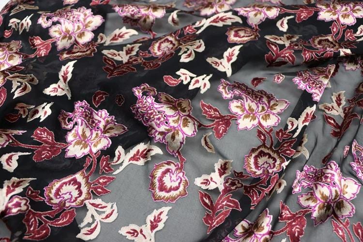a close-up view of a fabric with a floral pattern in shades of black, pink, and white. The fabric appears to be made of silk or a similar material, and the pattern consists of various flowers and leaves in different sizes and colors. The fabric is crumpled, giving it a textured appearance.