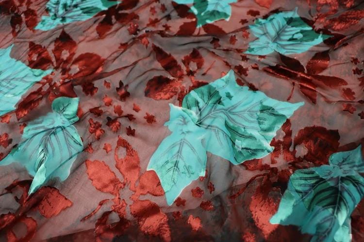 a close-up view of a fabric with a floral pattern. The fabric has a mix of red and green colors, with some areas appearing to be red and others green. The pattern consists of various leaf shapes and designs, creating a visually interesting and textured appearance.
