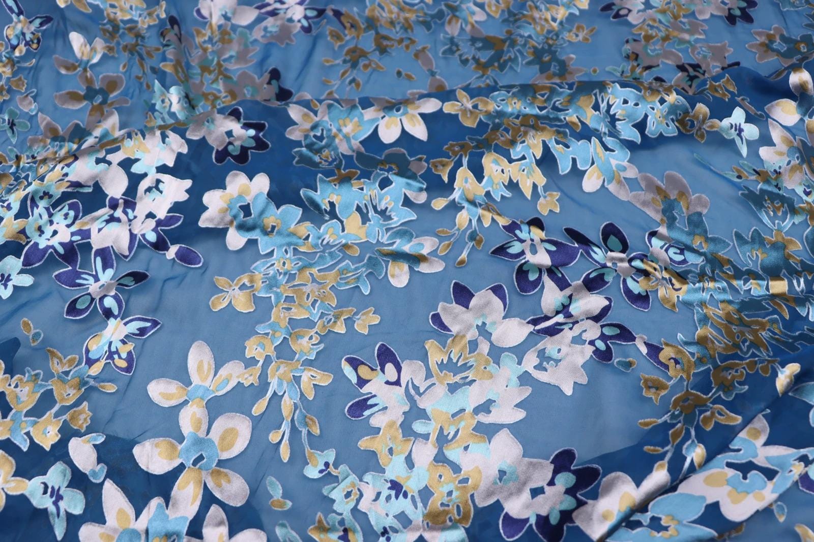 a close-up view of a fabric with a floral pattern in shades of blue and gold. The fabric appears to be a sheer material, with a textured and wrinkled appearance.
