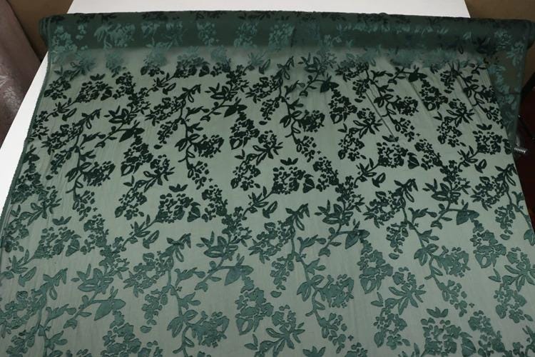 a close-up view of a fabric with a floral pattern, featuring green leaves and flowers. The fabric appears to be a type of lace or sheer material, with a repeating pattern of green leaves and flowers across the surface.