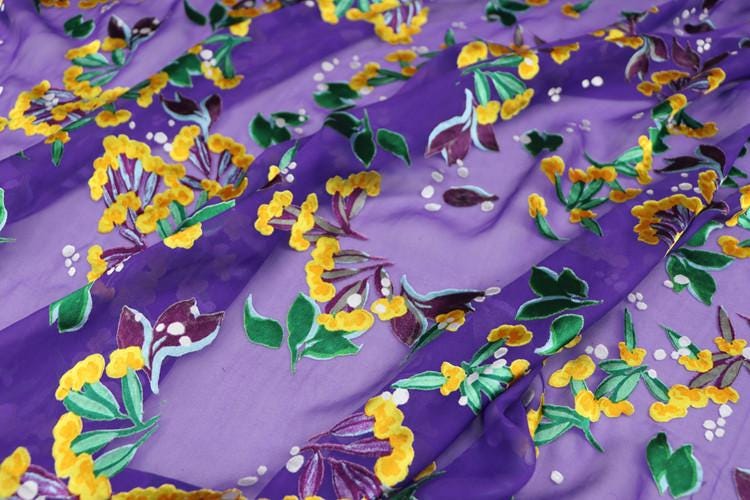 a close-up view of a fabric with a vibrant floral pattern in shades of purple and yellow. The pattern features various flowers and leaves, creating a visually striking and colorful design.