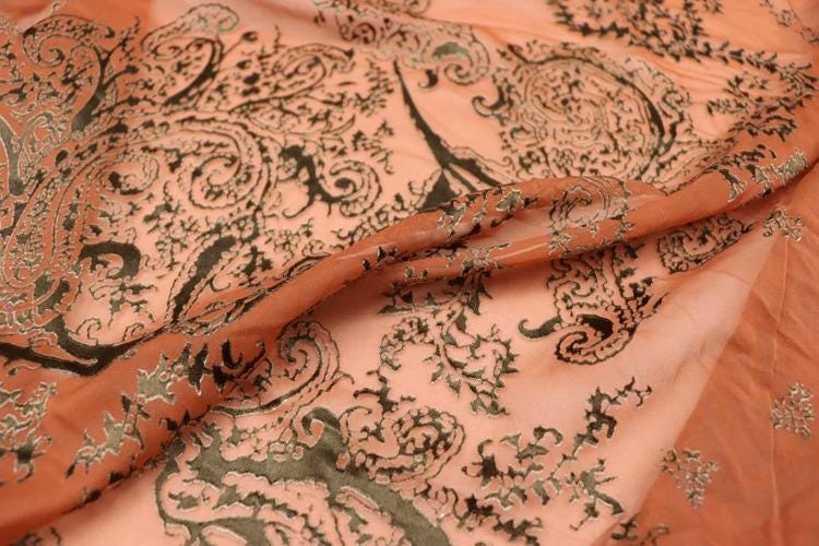 a close-up view of a fabric with a paisley pattern in shades of orange and black. The fabric appears to be made of silk or a similar material, and the intricate design of the paisley pattern is clearly visible.
