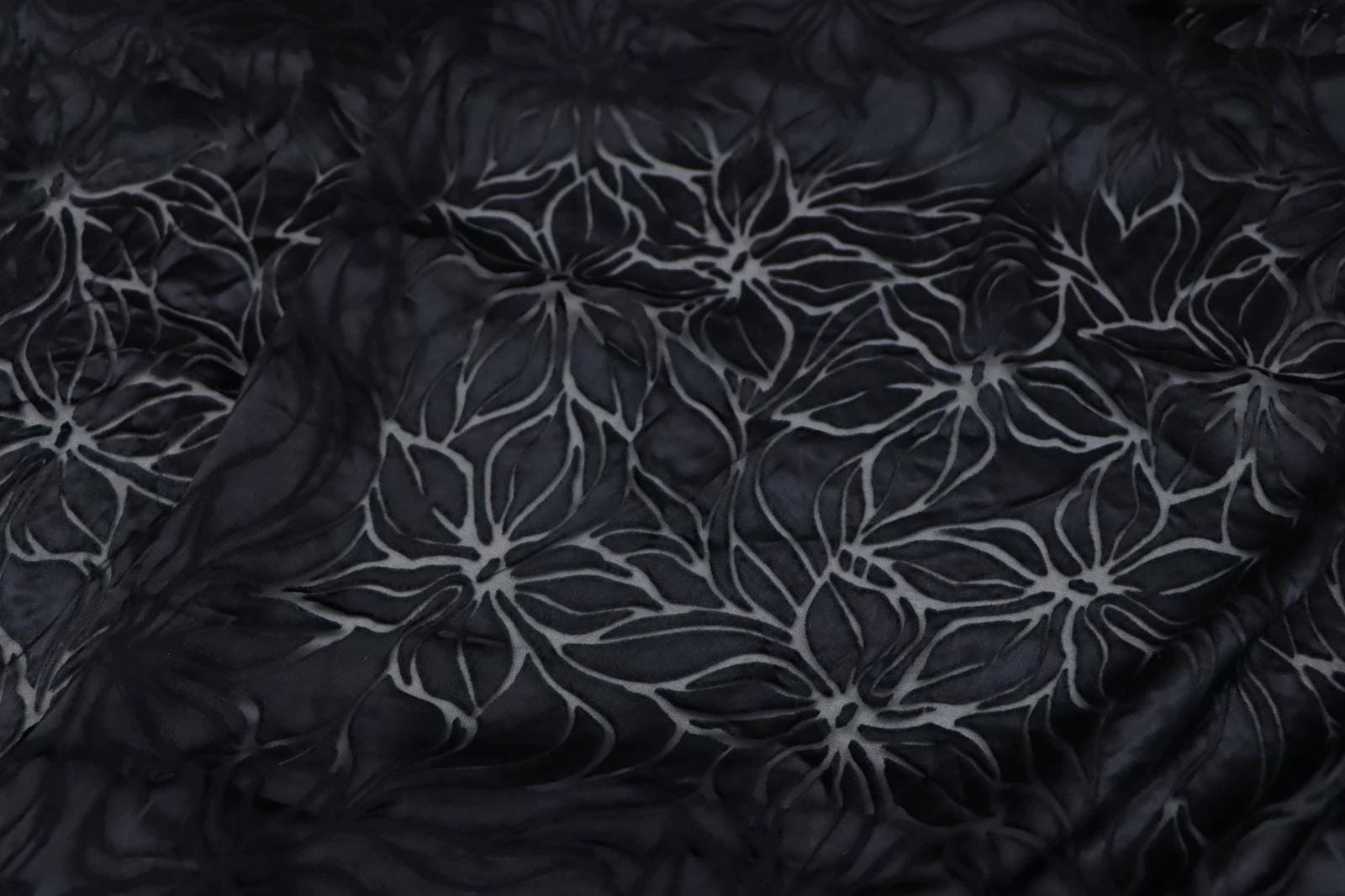 a close-up view of a dark fabric with a repeating floral pattern. The pattern consists of stylized white flowers or leaves against a black background, creating a visually striking contrast.