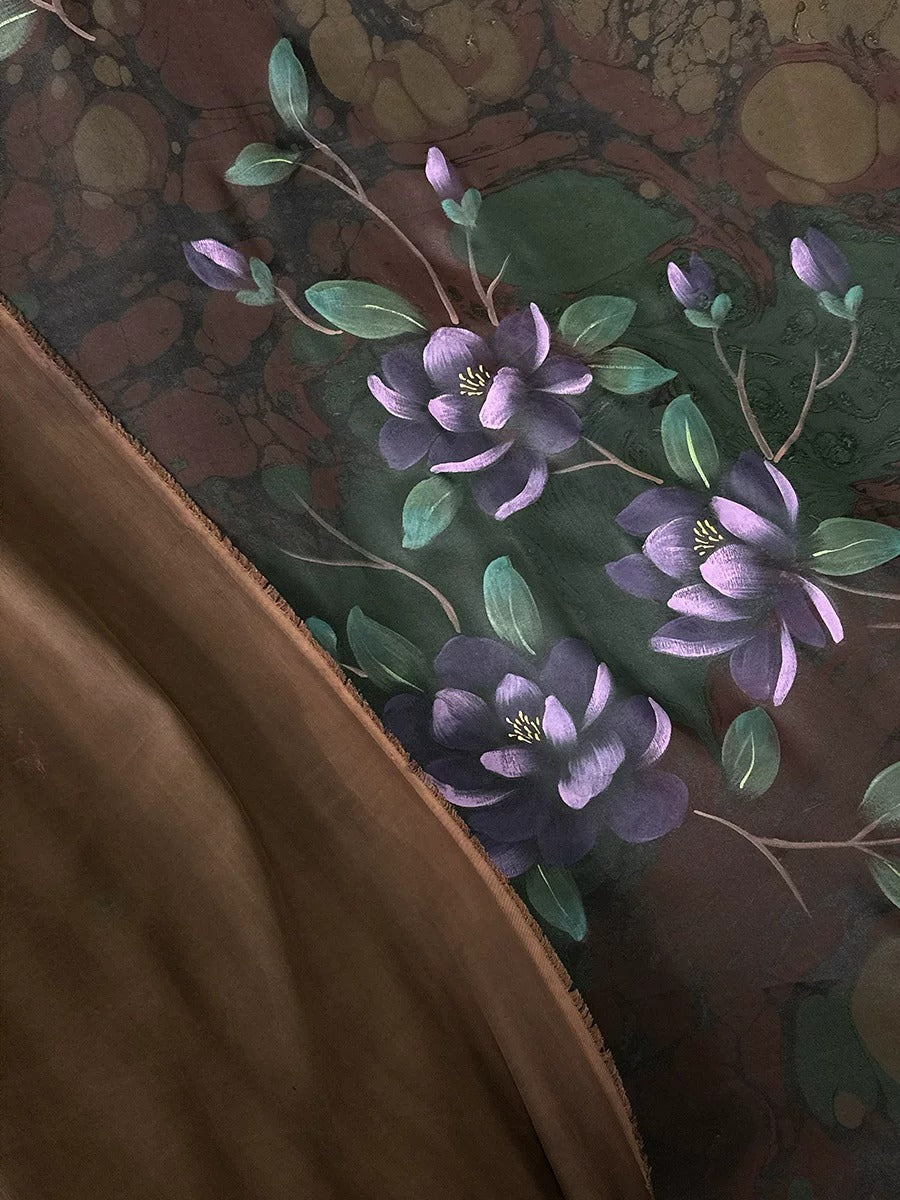 a close-up view of a floral pattern on a fabric, with purple flowers and green leaves. The background appears to be a dark brown color, creating a contrast with the vibrant colors of the flowers.