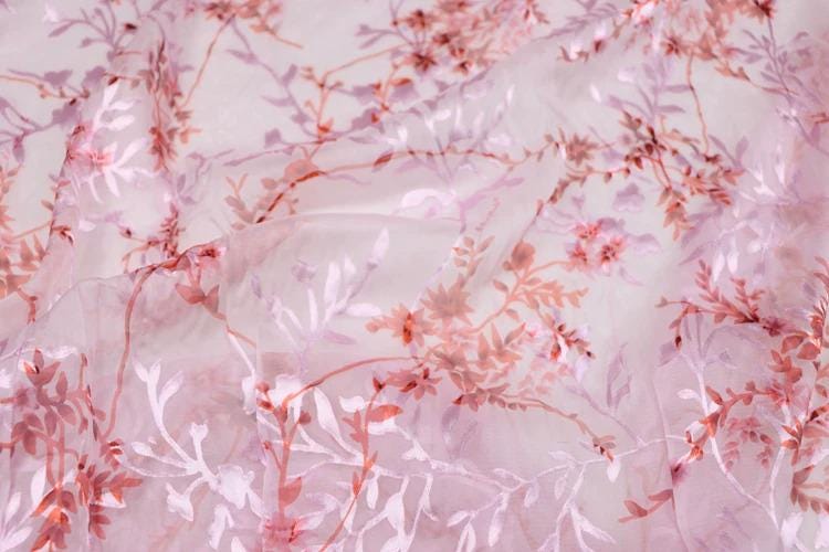 a close-up view of a fabric with a pink and white floral pattern. The fabric appears to be made of sheer material, with delicate pink and white flowers and leaves visible on the surface. The pattern is intricate and detailed, creating a visually appealing and textured appearance.